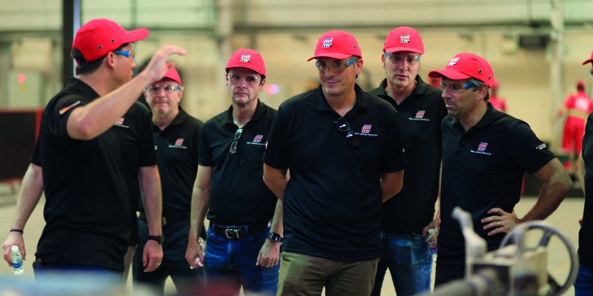 Gruppe von Personen mit roten Hüten und schwarzen Shirts, die einer Präsentation in einer Industrieumgebung zuhören.